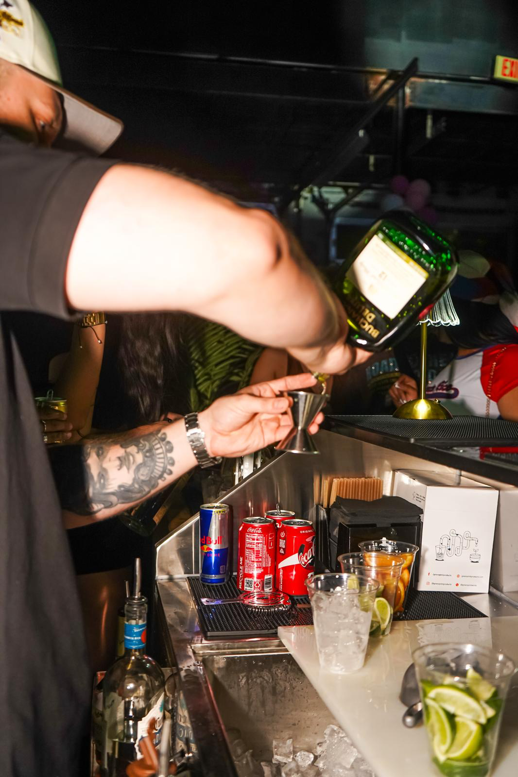 Bartender pouring a shot of herbal liqueur into a jigger behind the bar at a Los Primos event